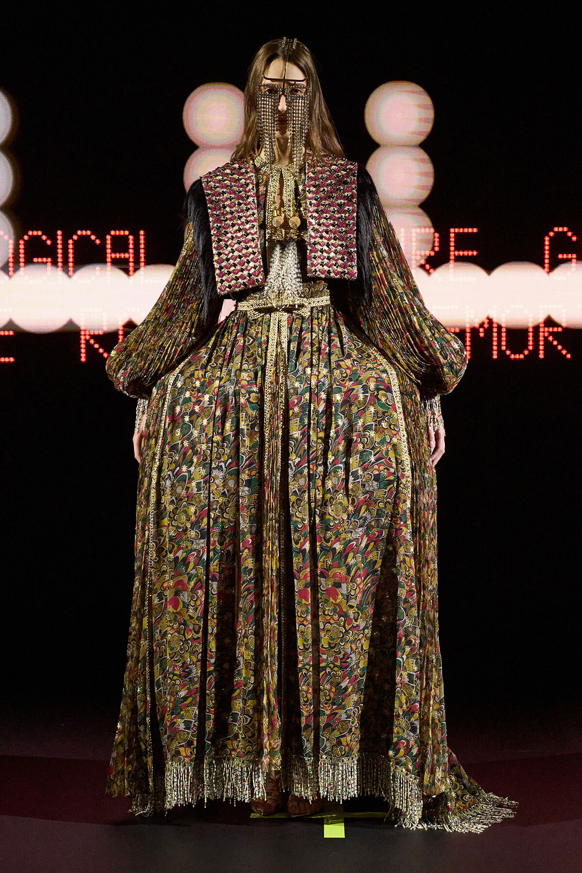 Model on a runway wearing an ornate, multicolored floral gown with gold fringe and beaded face veil.