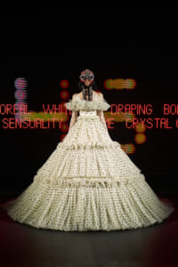 Model wearing a large off-white polka-dot ball gown with tiered ruffles and a jeweled masquerade mask on a dark runway backdrop with red neon text behind her.