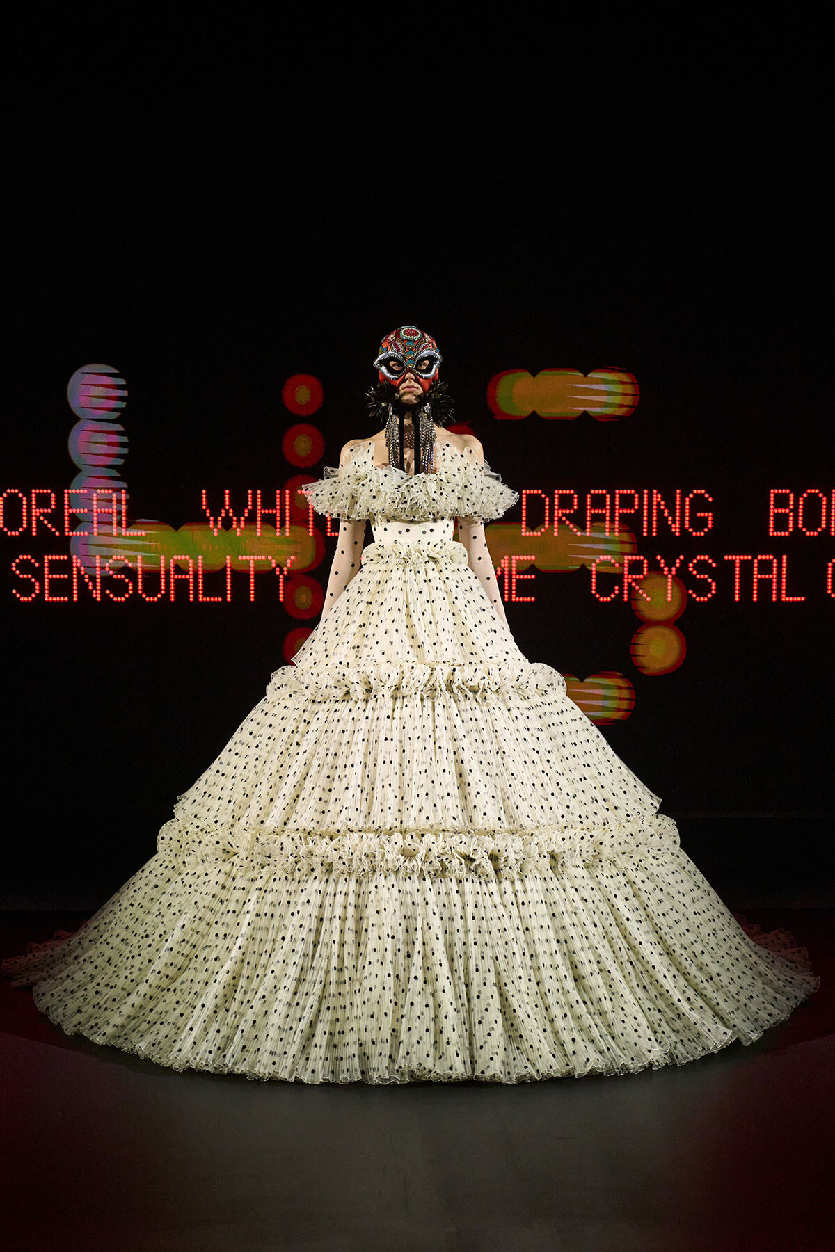 Model wearing a large off-white polka-dot ball gown with tiered ruffles and a jeweled masquerade mask on a dark runway backdrop with red neon text behind her.