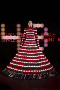 Model on a runway wearing a voluminous red, white, and black striped gown with fringe hem against a dark background with red LED text.