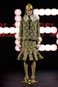 Fashion model on runway wearing an ornate gold-embellished suit with fur trim and a matching headdress, illuminated stage backdrop behind her.