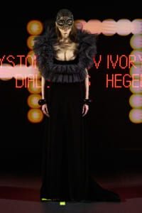 Model on a dark runway wearing a dramatic black gown with a feathered collar and lace mask, neon backdrop behind.