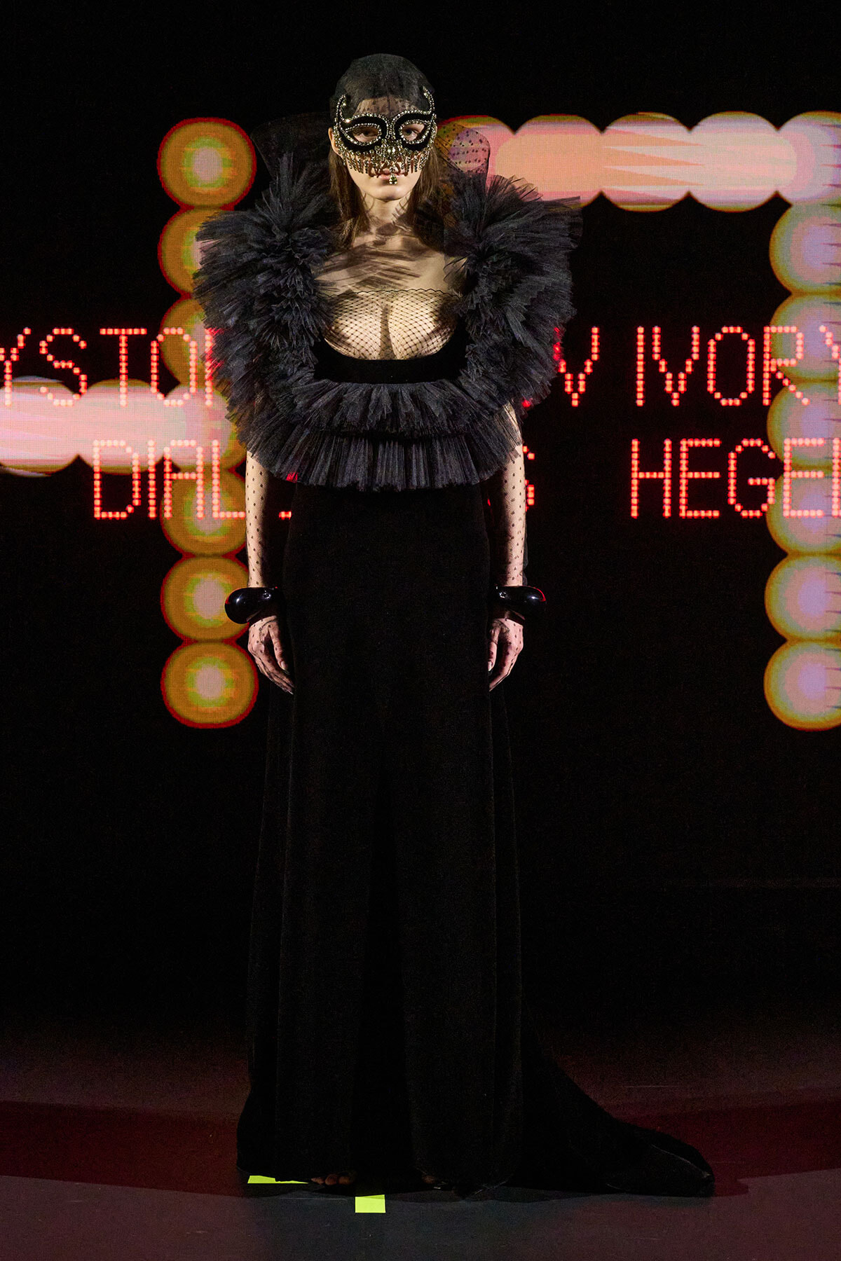 Model on a dark runway wearing a dramatic black gown with a feathered collar and lace mask, neon backdrop behind.