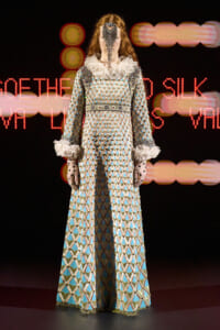 Model on a runway wearing a long, diamond-patterned gown with ruffled white cuffs and a beaded face mask under stage lights.