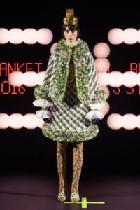Runway model in a green and purple diamond-patterned coat with a fluffy scarf, ruffled white cuffs, gold headpiece, and lace tights.