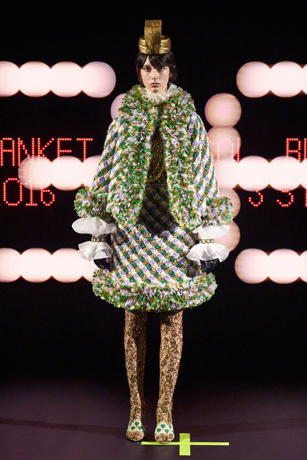 Runway model in a green and purple diamond-patterned coat with a fluffy scarf, ruffled white cuffs, gold headpiece, and lace tights.