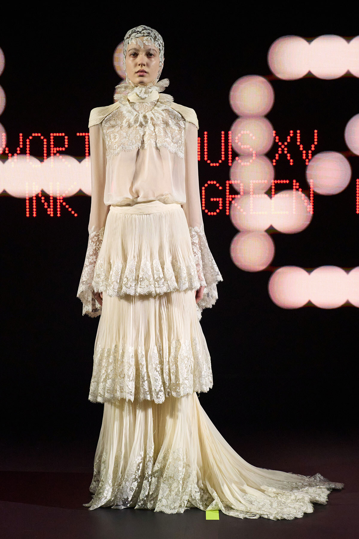 Model on a runway wearing an ivory lace gown with high neck and layered ruffled skirt, train trailing behind.