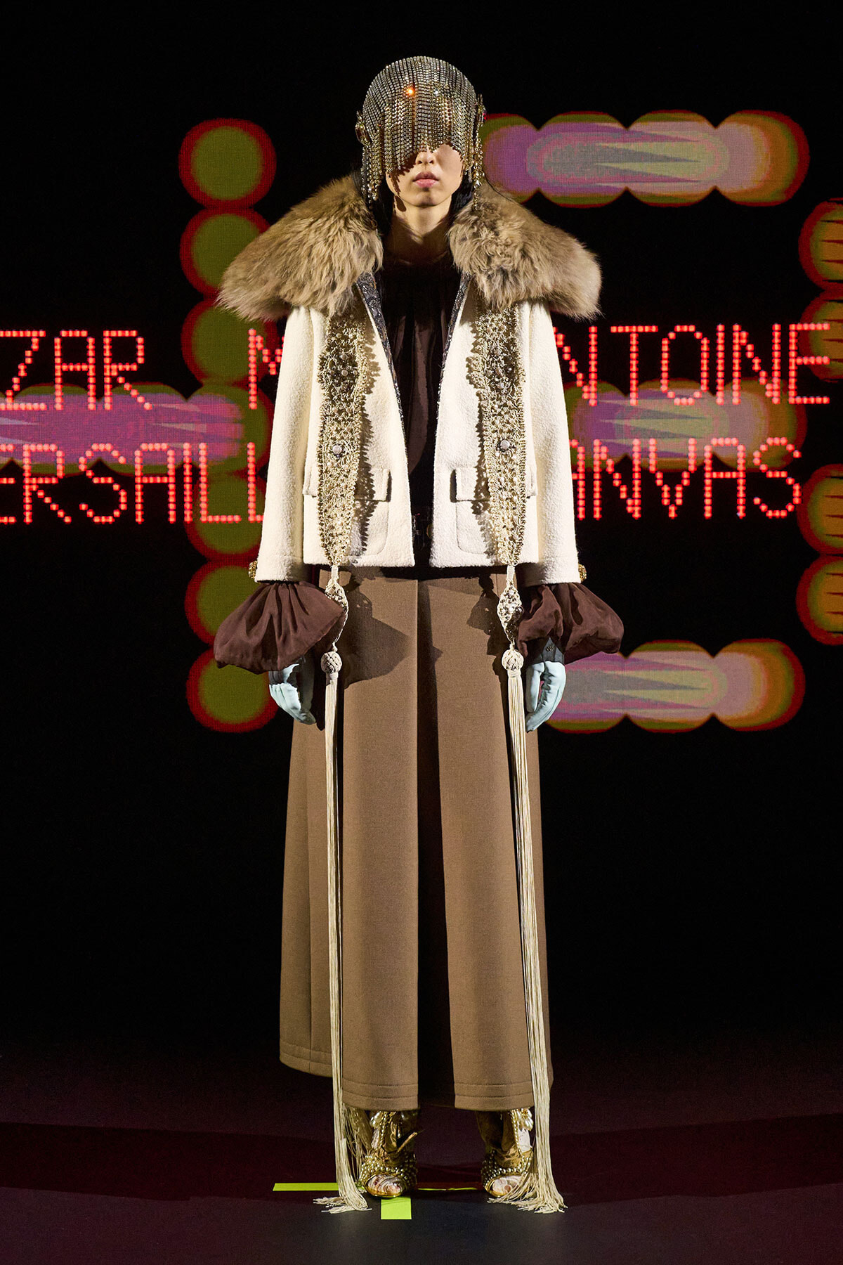 Runway model wearing a white fur-trimmed coat with a jeweled headpiece and beige trousers, holding two ceremonial swords.