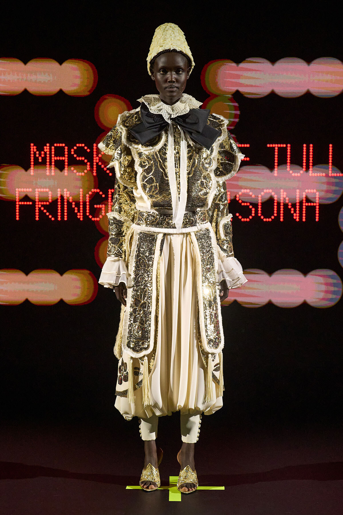 Model on a runway wearing an ornate gold-embellished ensemble with lace, ruffles, and a tall yellow headdress; neon stage lights in the background.