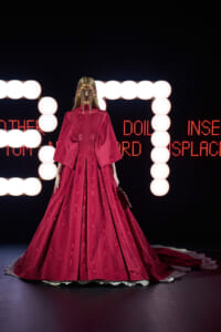 Model in a long, bright pink ballgown with dramatic sleeves on a runway, ornate chain mask, neon orbs behind and red LED text.