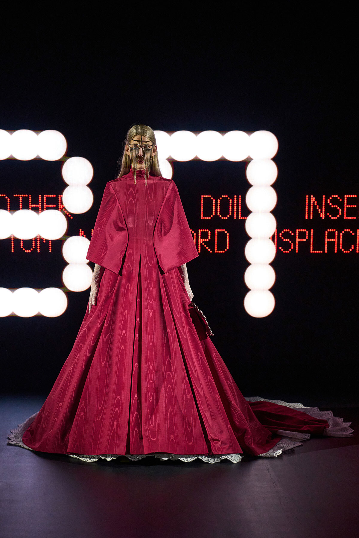 Model in a long, bright pink ballgown with dramatic sleeves on a runway, ornate chain mask, neon orbs behind and red LED text.