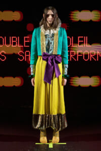 Model standing on a runway in a bright teal jacket, yellow skirt, and a large purple bow belt, with lace collar and metallic hems, against a neon red background.