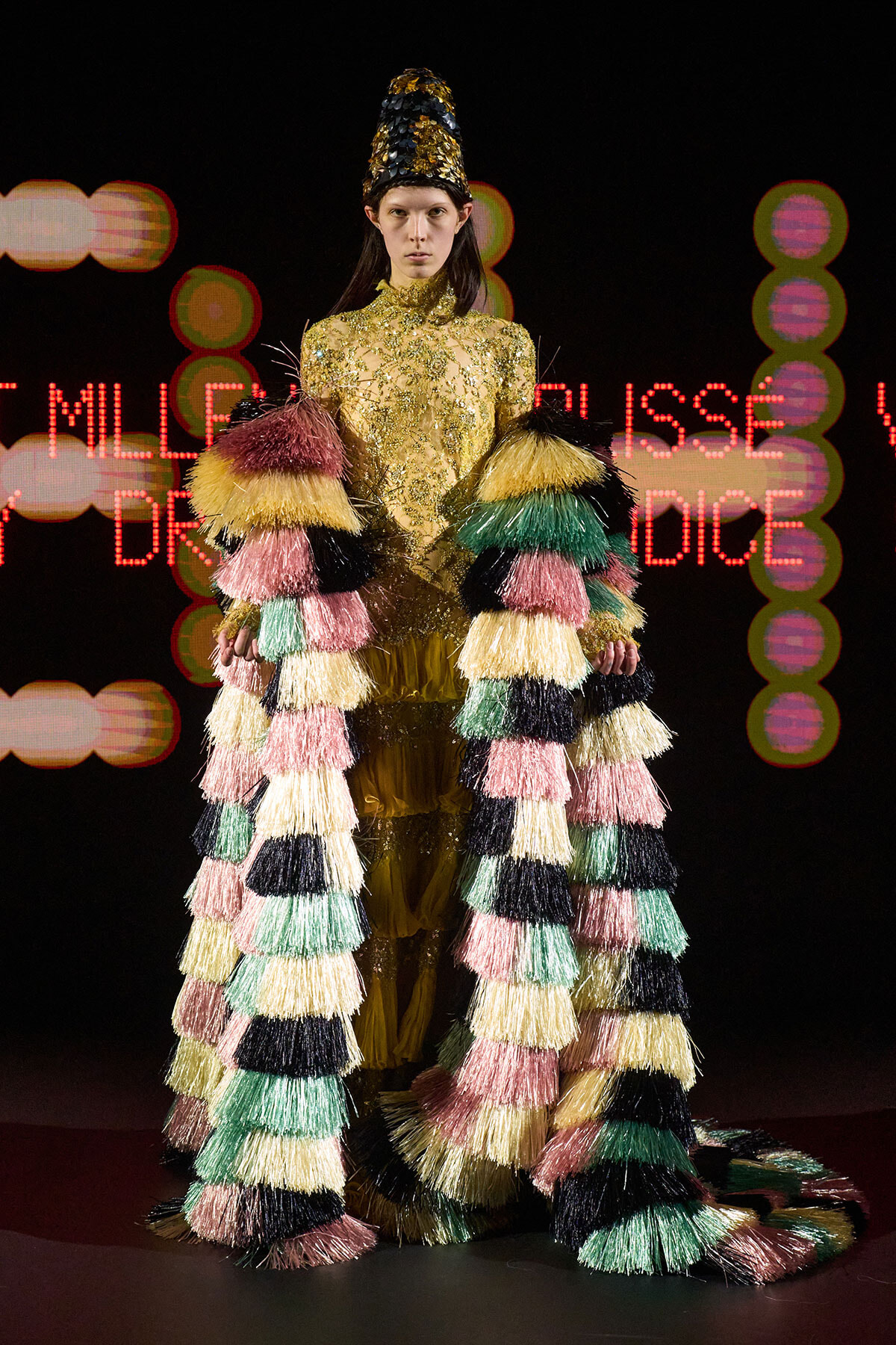 Model in a gold sequined dress with a long multicolored fringe cape on a runway with neon lights in the background.
