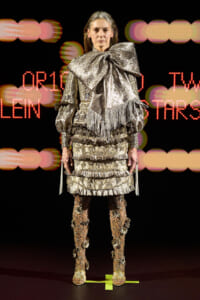 Model on a runway wearing an ornate metallic silver fringe outfit with a large bow and embellished boots, against a neon background.