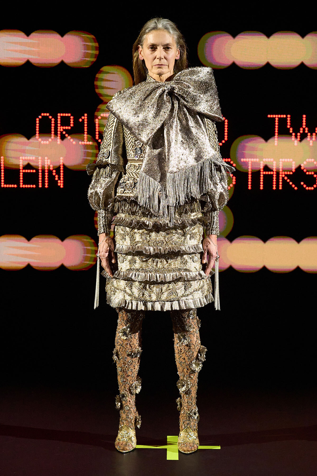 Model on a runway wearing an ornate metallic silver fringe outfit with a large bow and embellished boots, against a neon background.