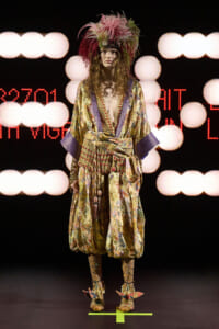 Model wearing a gold floral kimono with purple cuffs and a dramatic feathered headpiece on a runway stage in bright lights, front view.