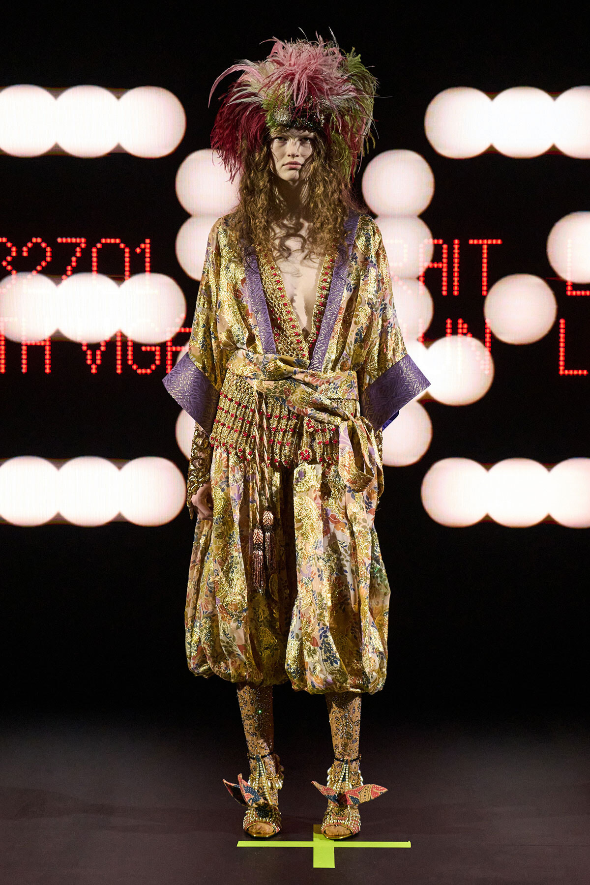 Model wearing a gold floral kimono with purple cuffs and a dramatic feathered headpiece on a runway stage in bright lights, front view.