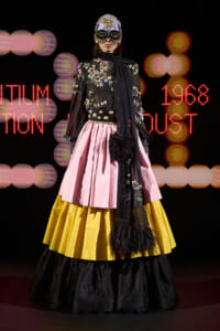 Model on runway wearing a black floral embroidered top with a pink and yellow tiered skirt and a jeweled mask, against a neon backdrop