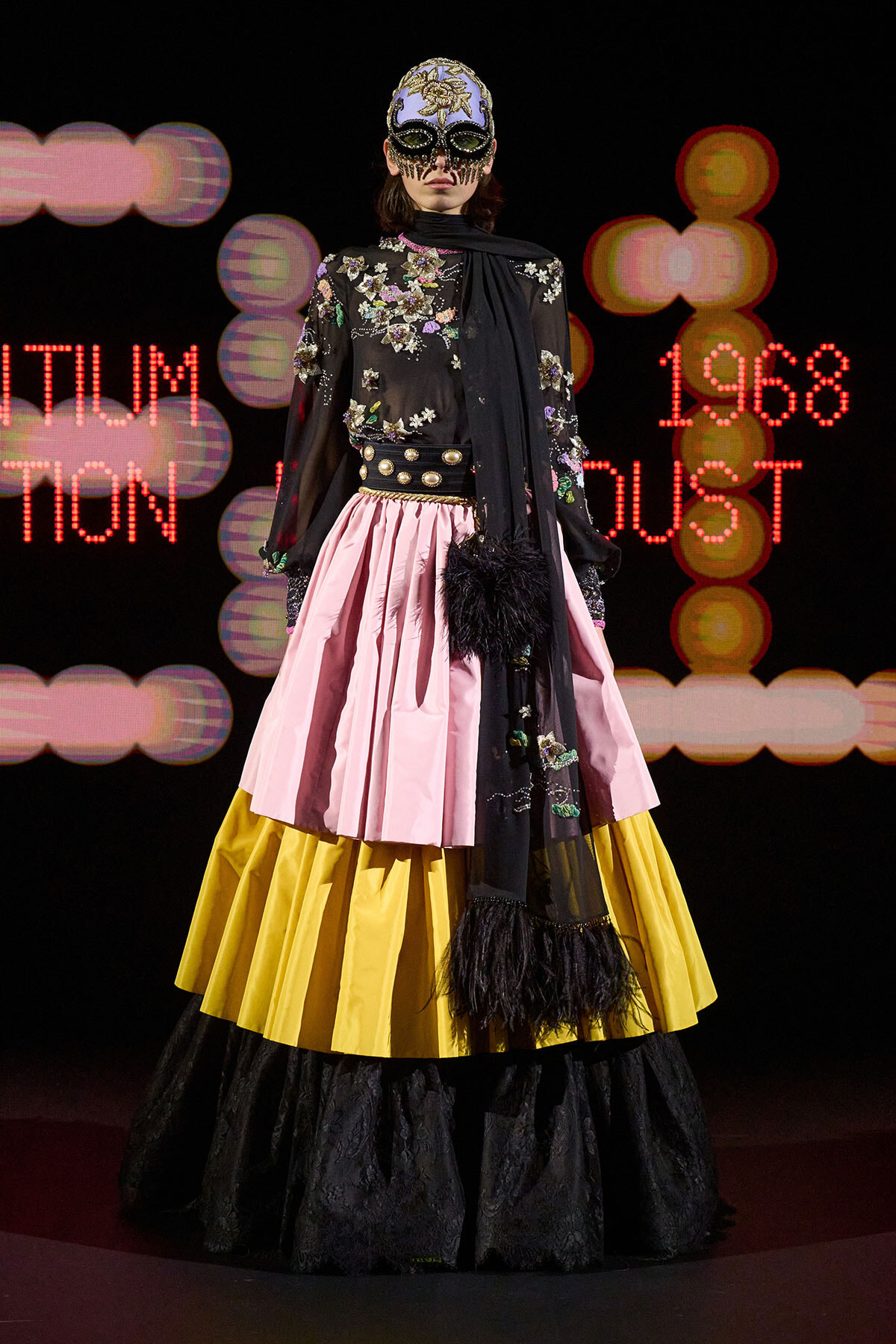 Model on runway wearing a black floral embroidered top with a pink and yellow tiered skirt and a jeweled mask, against a neon backdrop