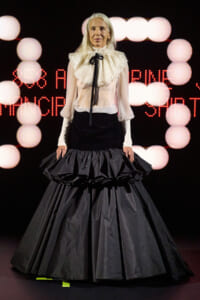 Older woman on stage wearing a white ruffled blouse with black ribbon and a voluminous black ball gown with tiered ruffles, against a backdrop of pink stage lights.