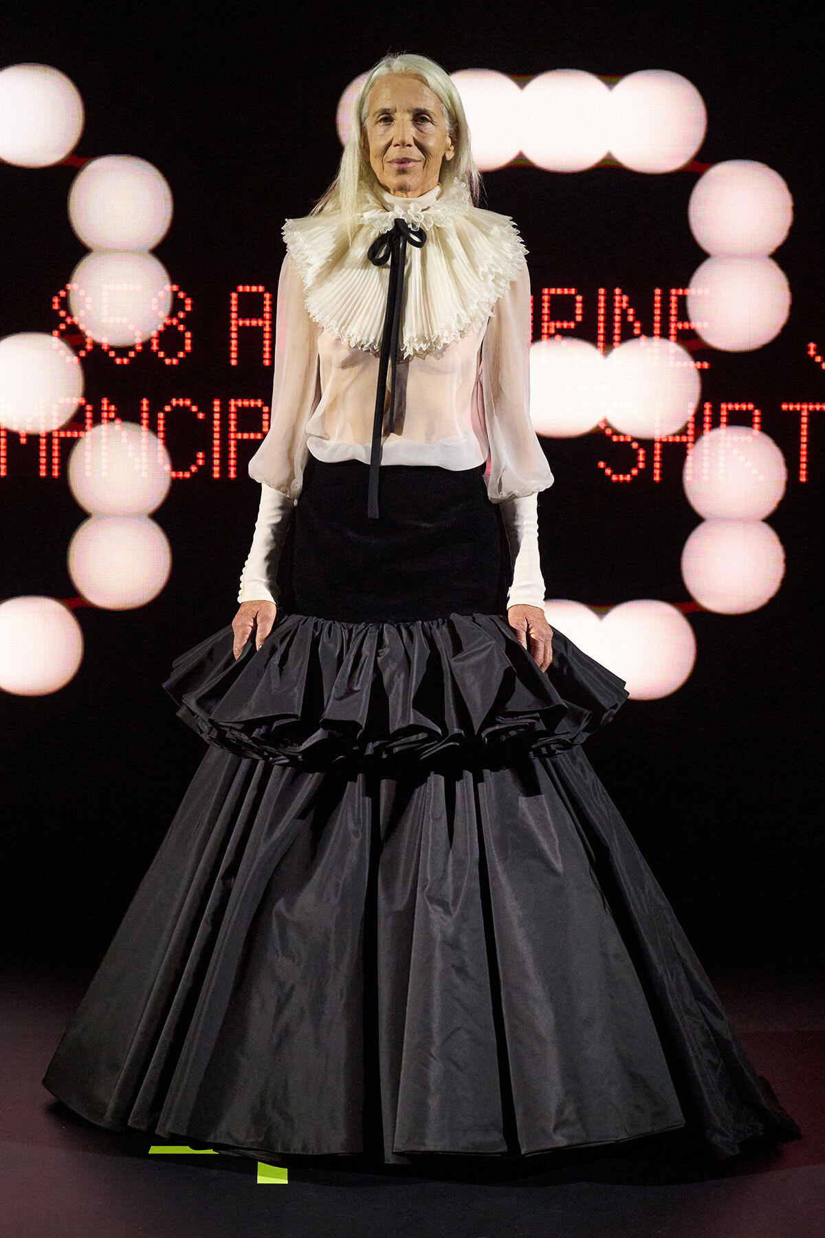Older woman on stage wearing a white ruffled blouse with black ribbon and a voluminous black ball gown with tiered ruffles, against a backdrop of pink stage lights.