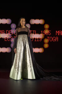 Female model on a runway wearing a black sheer-top gown with a long, sequined striped skirt, backdrop of red LED text.