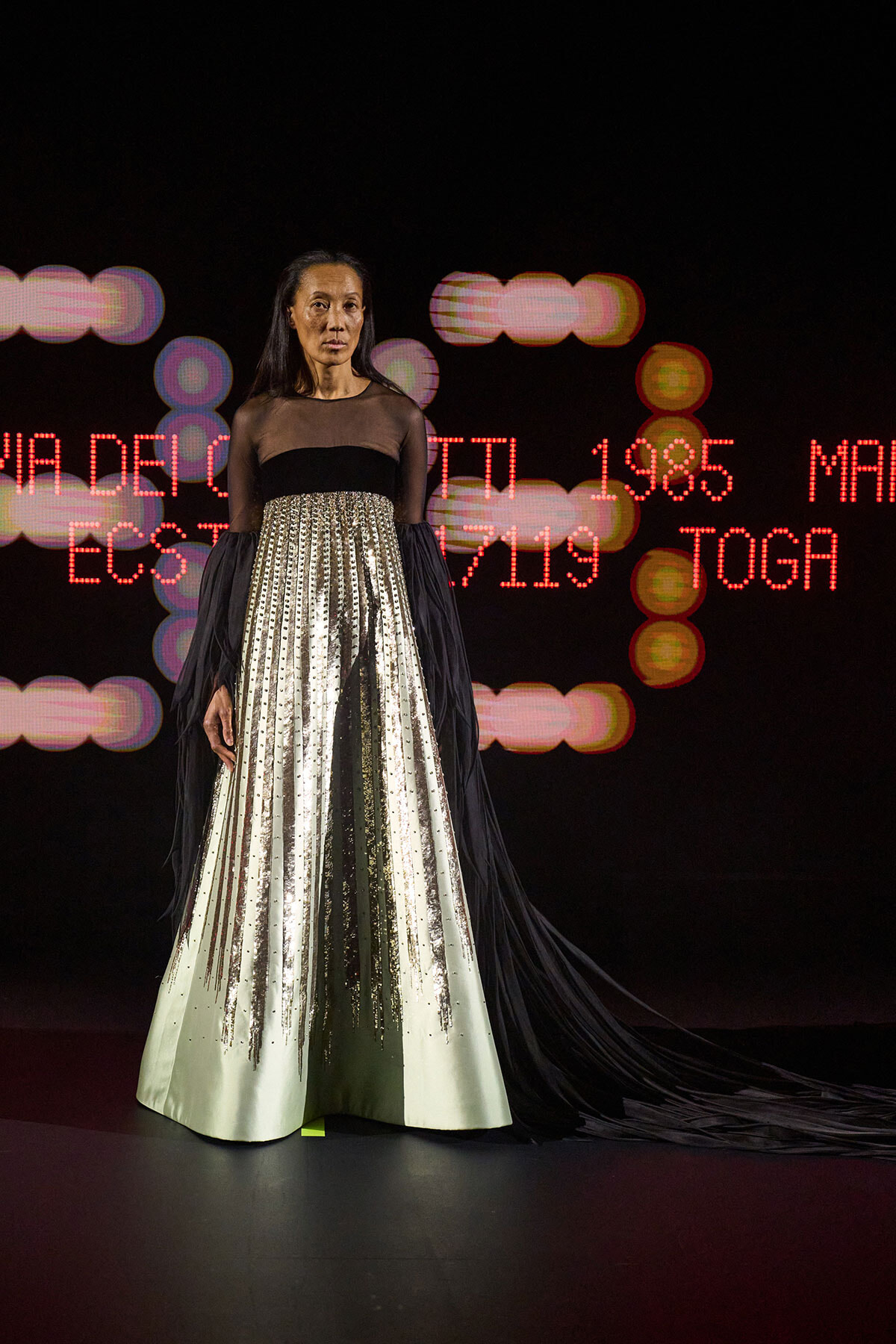 Female model on a runway wearing a black sheer-top gown with a long, sequined striped skirt, backdrop of red LED text.
