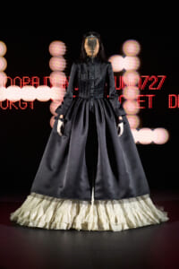 Model in a black Victorian-inspired gown with a voluminous skirt and cream ruffled hem, wearing a jeweled chain headpiece on a fashion show runway with red LED lights behind.