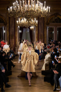 Model walks a fashion runway in a grand, chandelier-lit ballroom wearing a beige oversized coat with a belt and large sleeves; photographers line the sides.