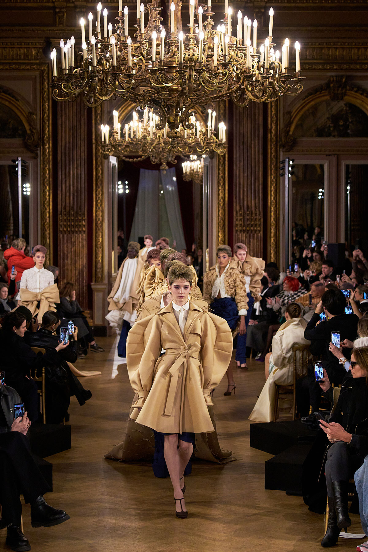 Model walks a fashion runway in a grand, chandelier-lit ballroom wearing a beige oversized coat with a belt and large sleeves; photographers line the sides.