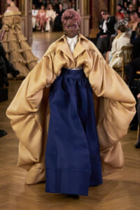 Runway model in oversized tan cape over a white shirt and navy skirt; audience watches from ornate room.