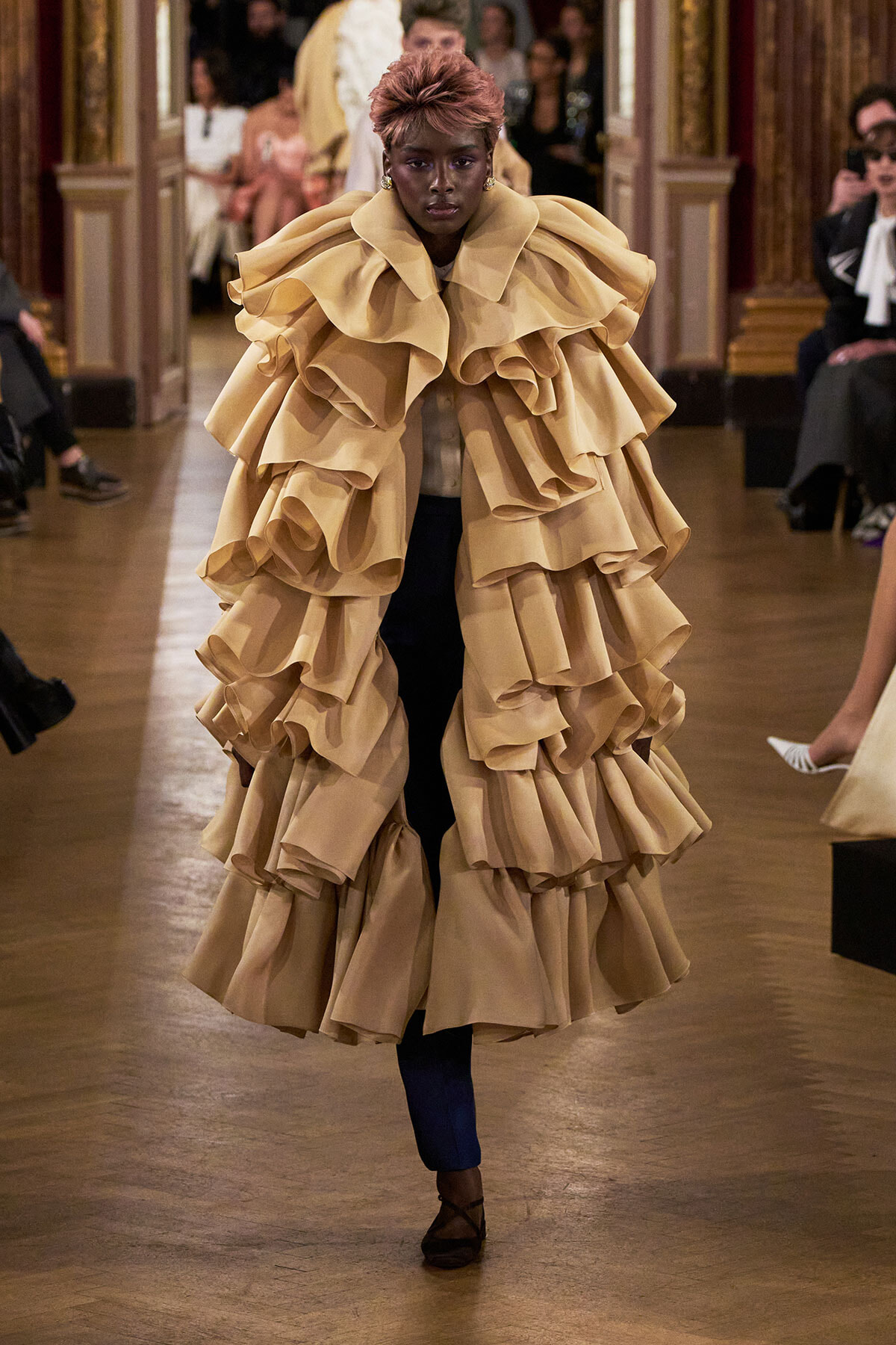Model walking the runway in a voluminous beige ruffle coat over blue pants, center-stage on a fashion show runway.