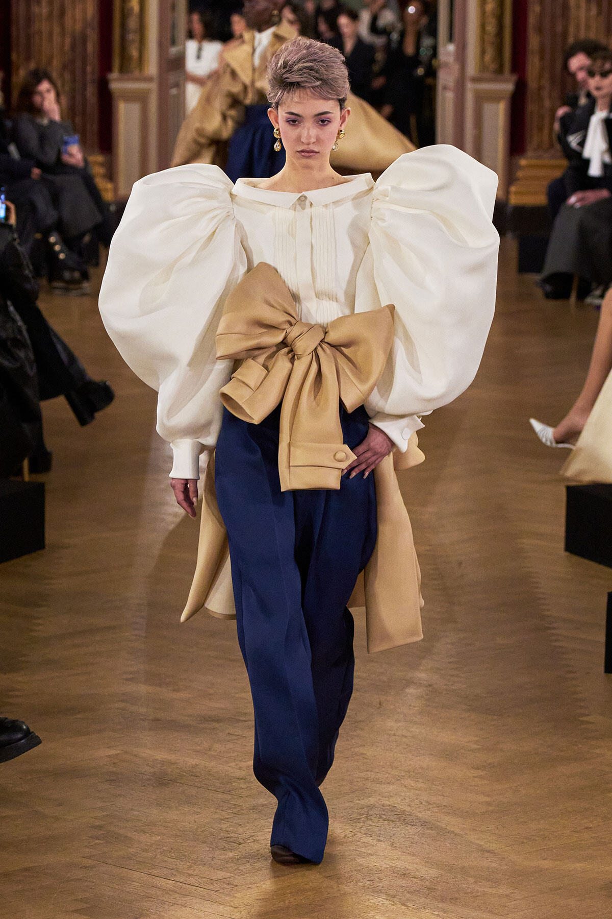 Model walking a runway in a white voluminous-sleeve blouse and navy trousers with a large beige bow at the waist