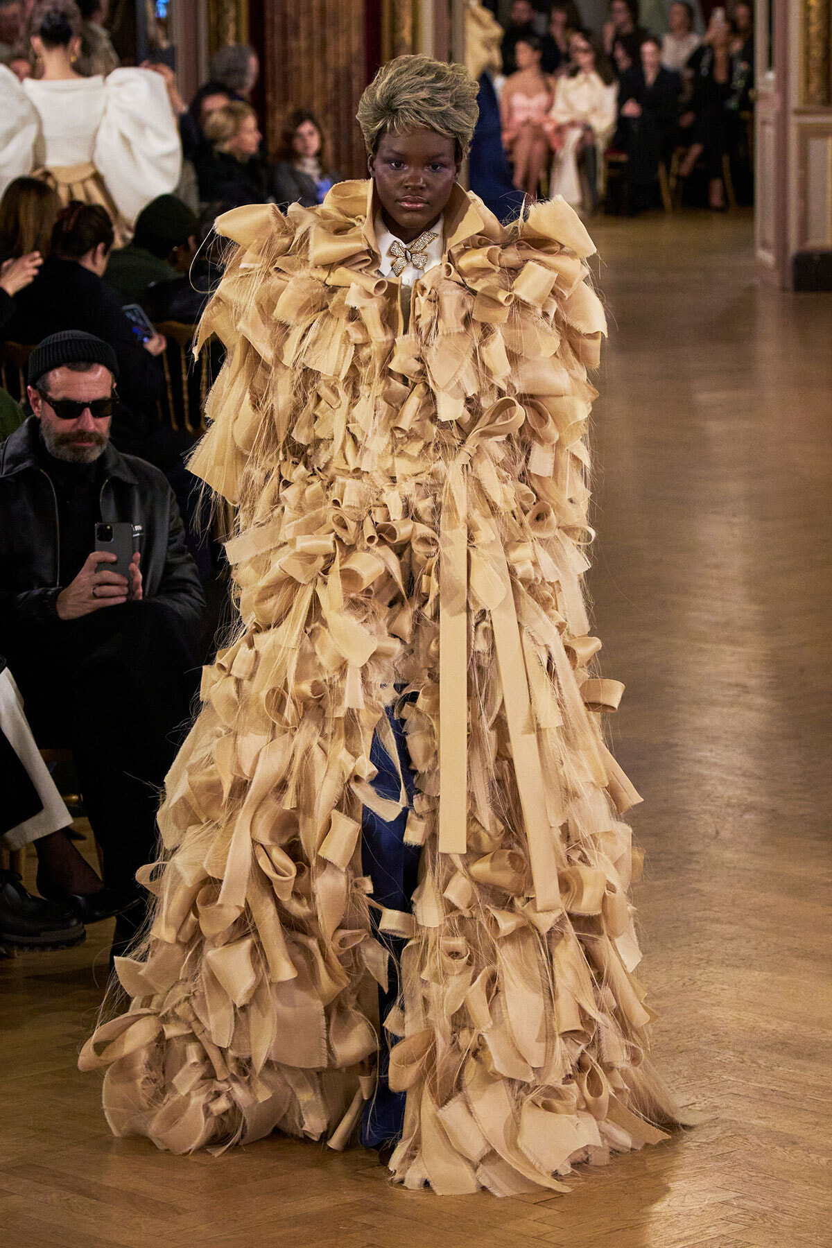 Model on a runway wearing an elaborate beige ruffled gown with fringe layers.
