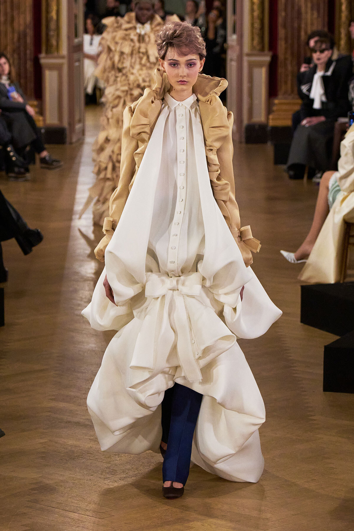 Model walking the runway in a voluminous white dress with a beige structured jacket over blue pants.