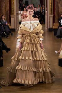 Model walks the runway in a voluminous beige tiered ruffled gown with off-the-shoulder white neckline, at a fashion show.