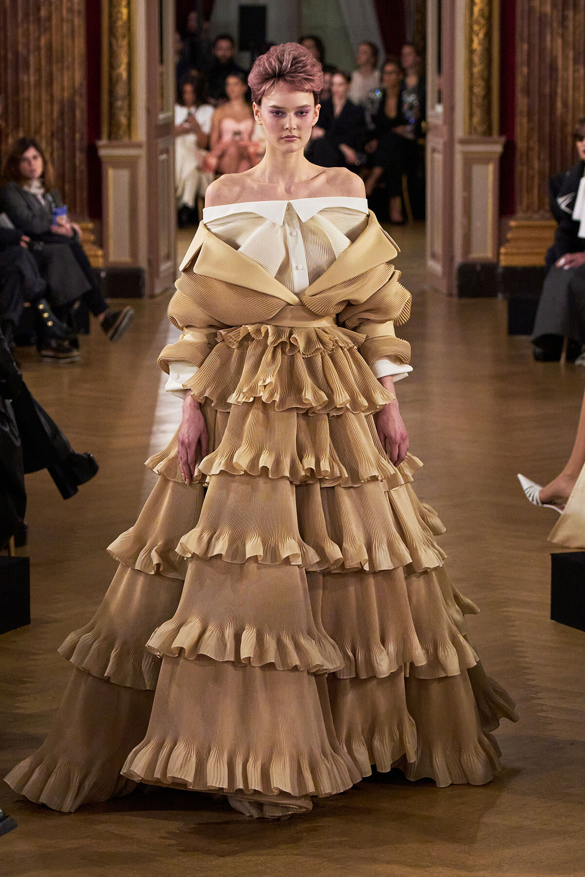 Model walks the runway in a voluminous beige tiered ruffled gown with off-the-shoulder white neckline, at a fashion show.