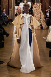 Model walking a fashion runway in a dramatic beige cape coat over a white blouse and asymmetric white and navy gown, hands on hips with an elegant stance.