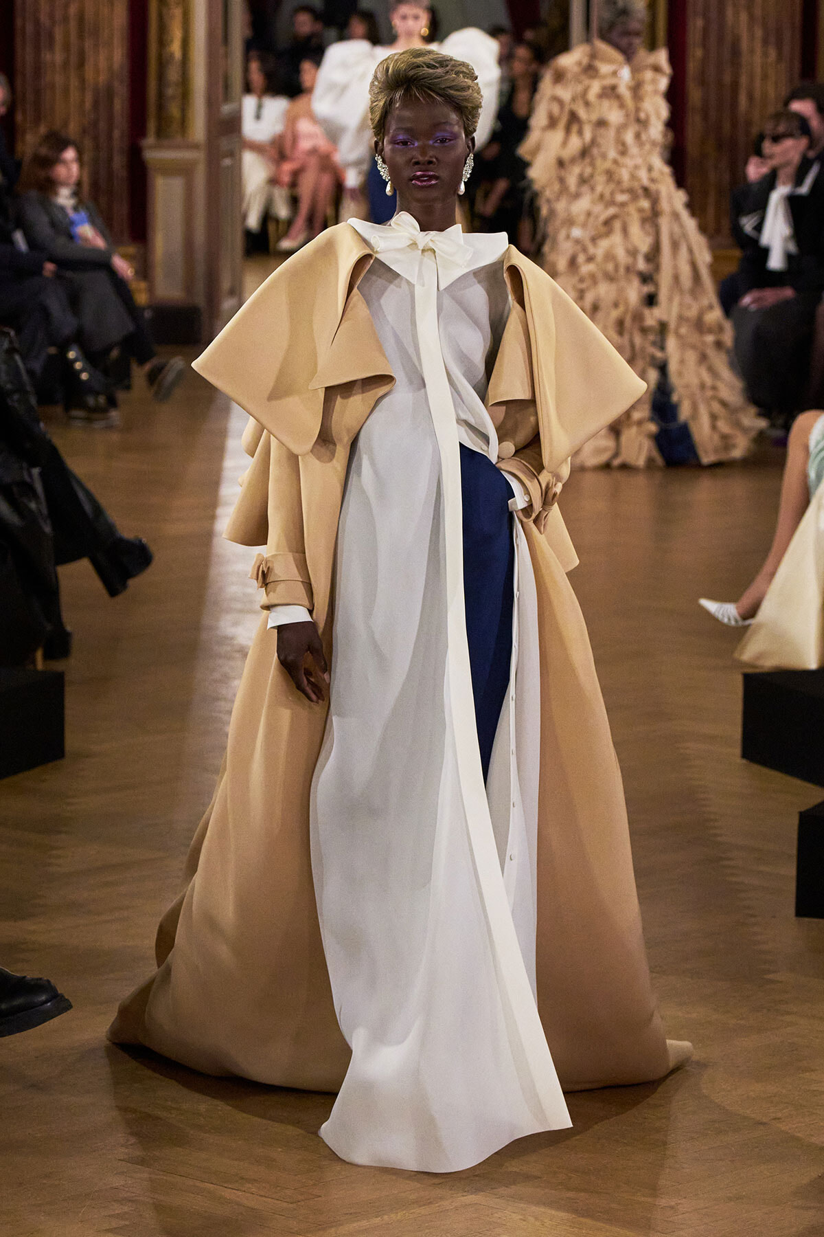 Model walking a fashion runway in a dramatic beige cape coat over a white blouse and asymmetric white and navy gown, hands on hips with an elegant stance.
