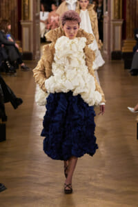 Model walks a fashion runway in a voluminous cream ruffle top and navy blue layered skirt with crinkled textures, mid-stride in a grand hall.