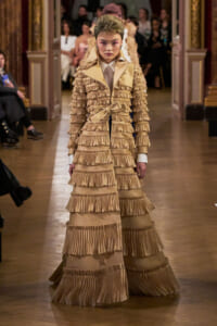 Model walking a fashion runway in a full-length beige gown with multiple tiers of pleated ruffles and a matching belt at the waist.
