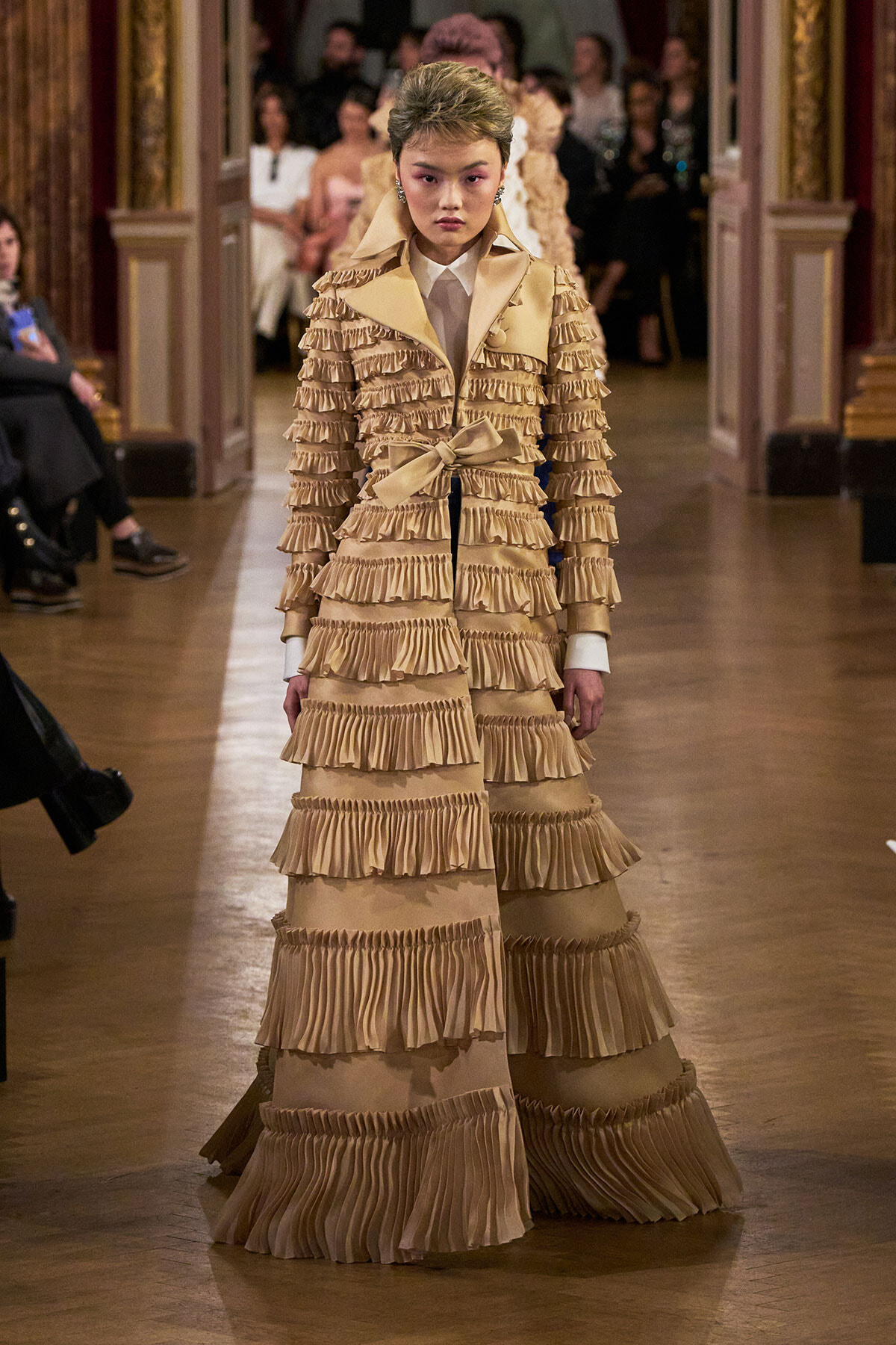 Model walking a fashion runway in a full-length beige gown with multiple tiers of pleated ruffles and a matching belt at the waist.