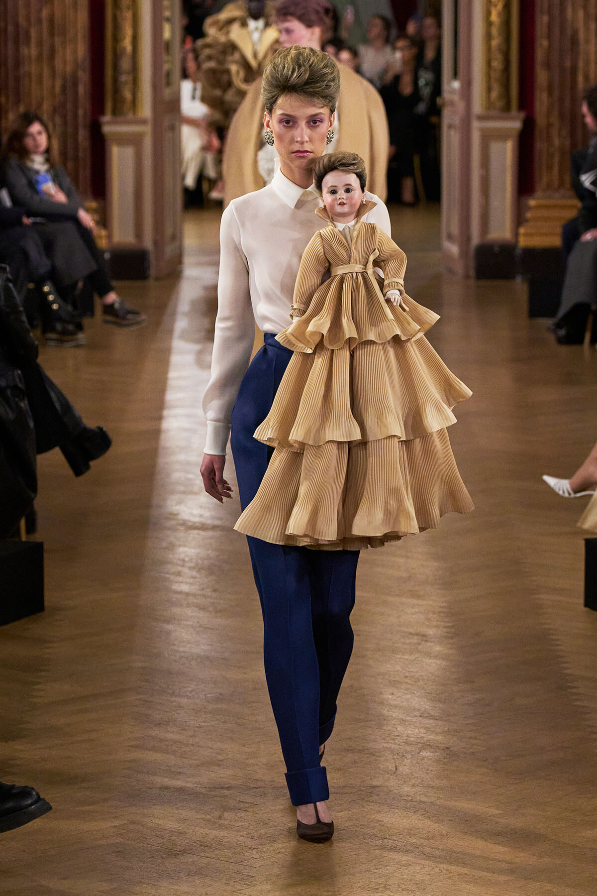 Model on a fashion runway carries a large vintage-style doll in a tan layered dress while audience watches in an ornate hall.