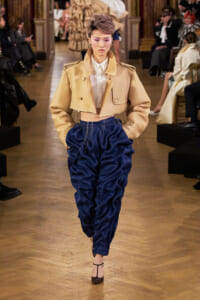 Model on runway wearing a beige cropped trench with oversized blue satin pants, audience in background.