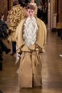 Model walks a fashion runway in an oversized beige trench coat with wide sleeves and belted waist, over a crinkled white top; ornate venue and audience blurred in the background.