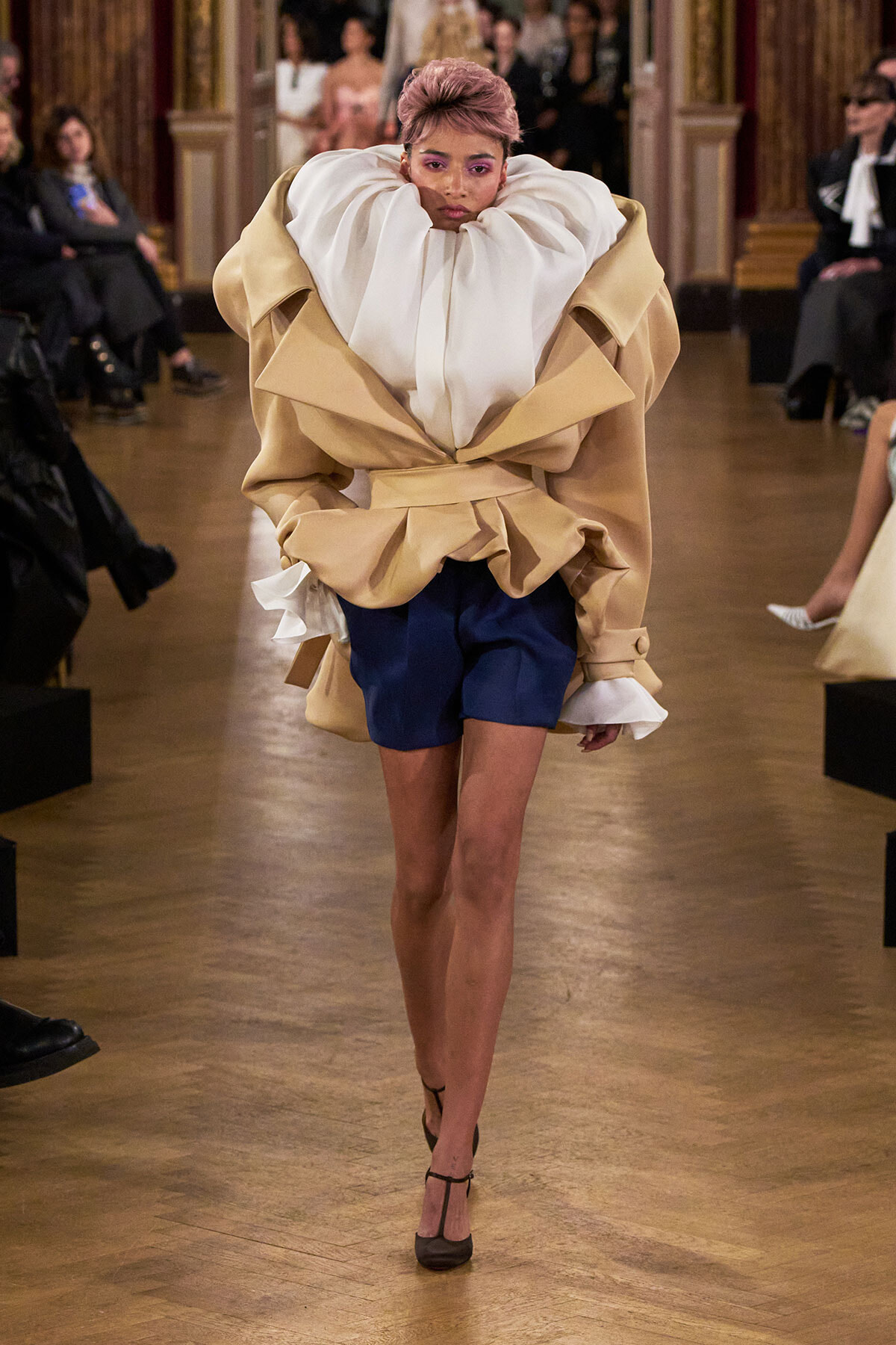 Model walking a fashion runway in a voluminous beige coat with a white ruffled high collar, navy shorts, and black heels, audience in the background.