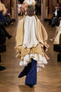 Model walking a fashion show in a voluminous cream cape over a white draped top and blue wide-leg pants.