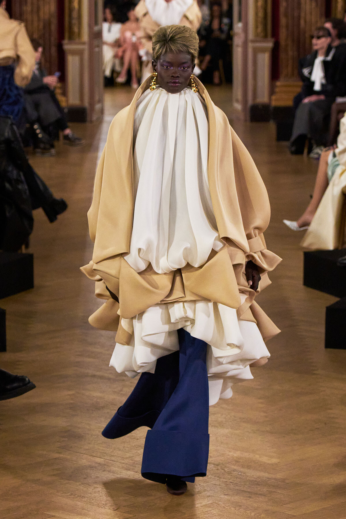 Model walking a fashion show in a voluminous cream cape over a white draped top and blue wide-leg pants.