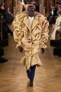 Model walks a runway in a voluminous beige coat with sculptural ruffles, paired with blue trousers and brown ankle boots.