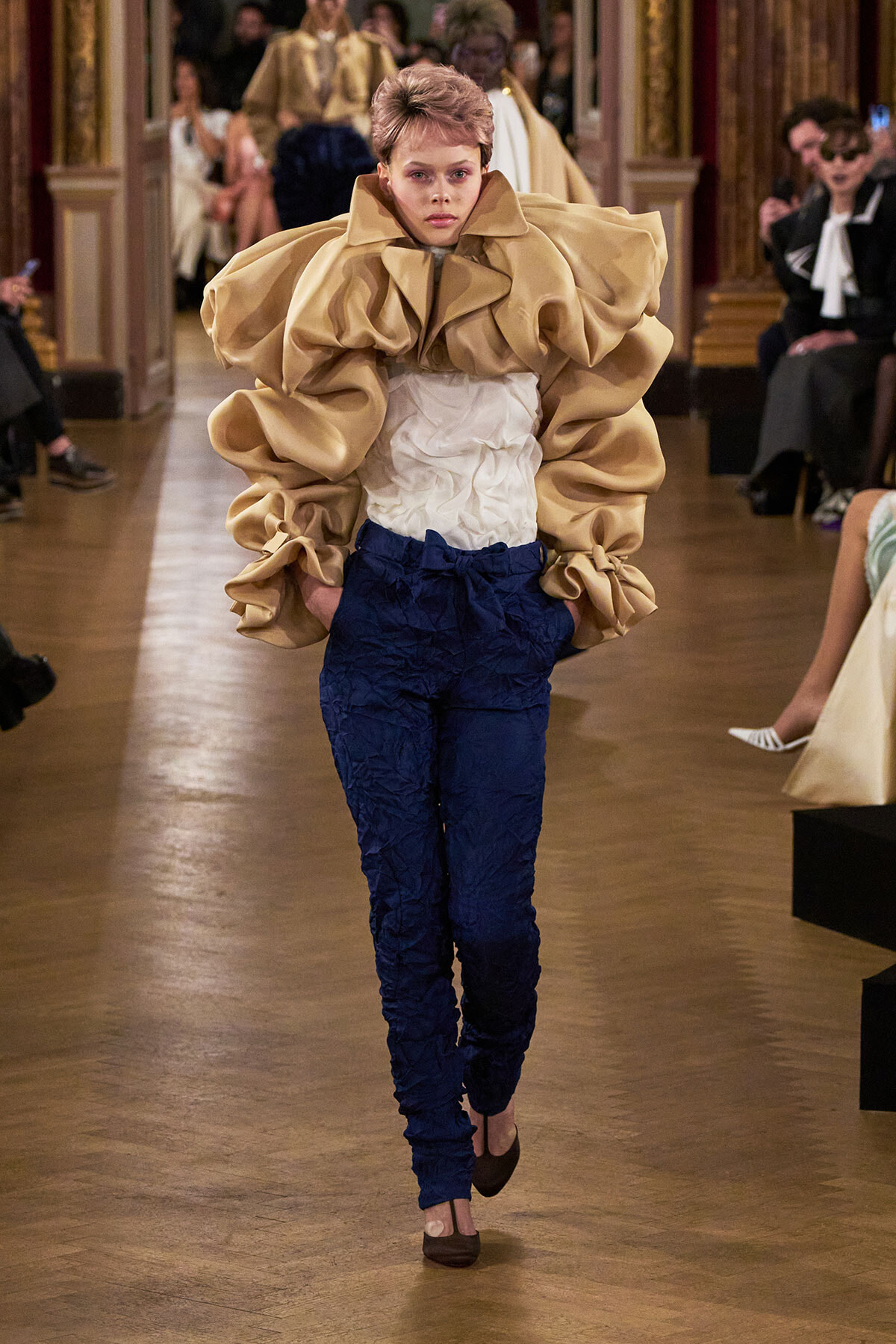 Model walking a runway in an oversized beige ruffled top and crinkled navy pants, white blouse peeking through, in a fashion show setting.